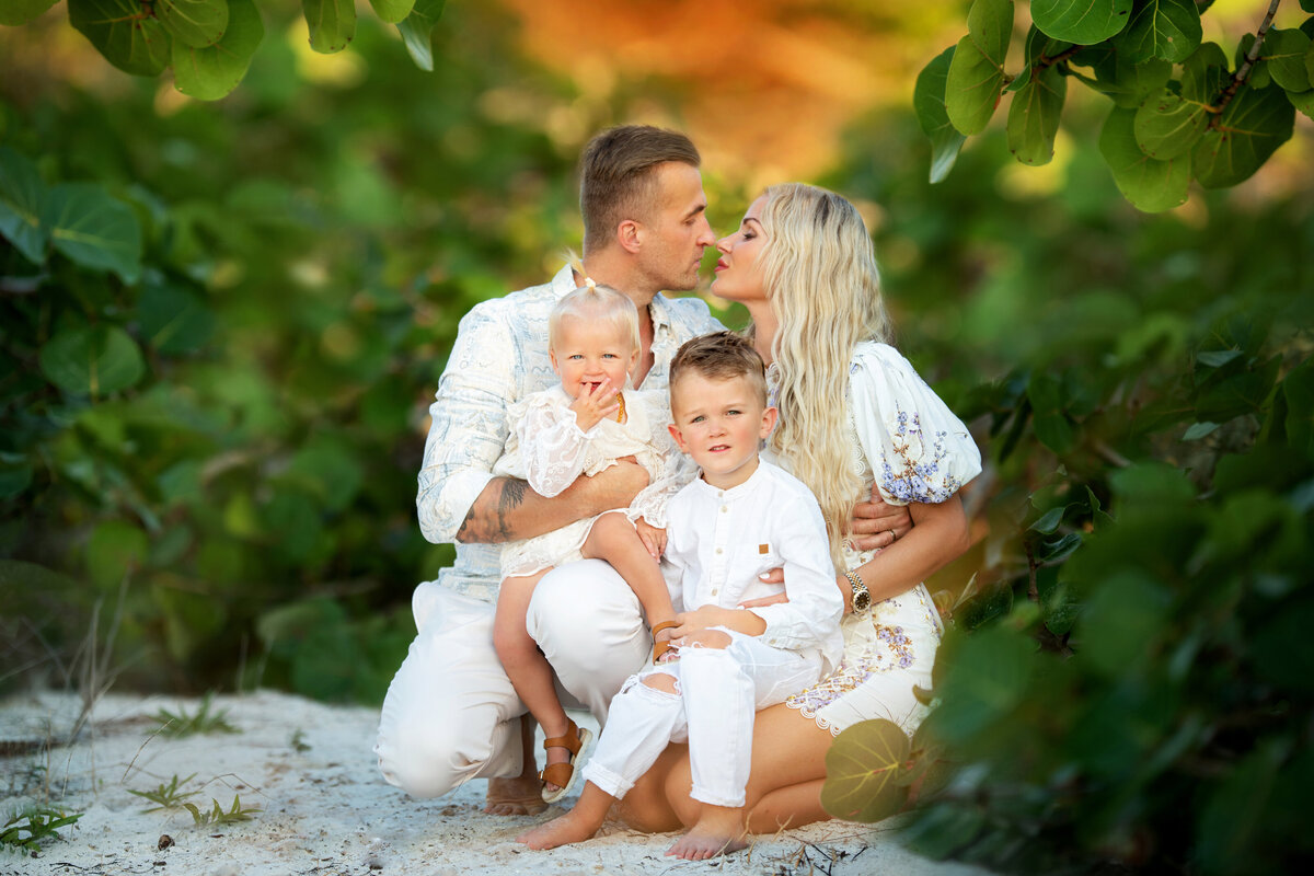Family dressed in white outfits sitting in the sand surrounded by green Florida leaves in Naples