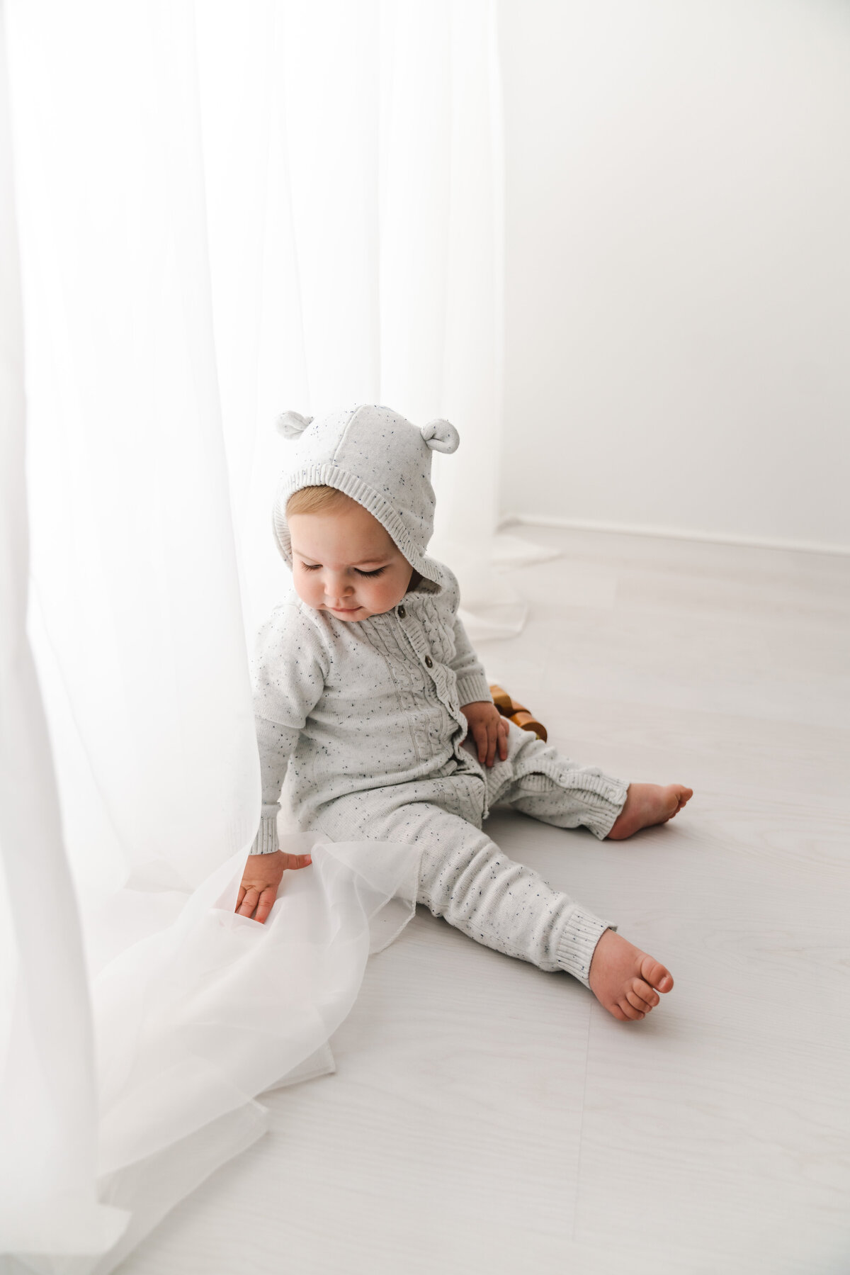 Toddler in cozy knitted outfit sitting on a white floor by a sheer curtain, with a contemplative expression. Taken by Fig and Olive Photography, Minneapolis Baby Photographer.