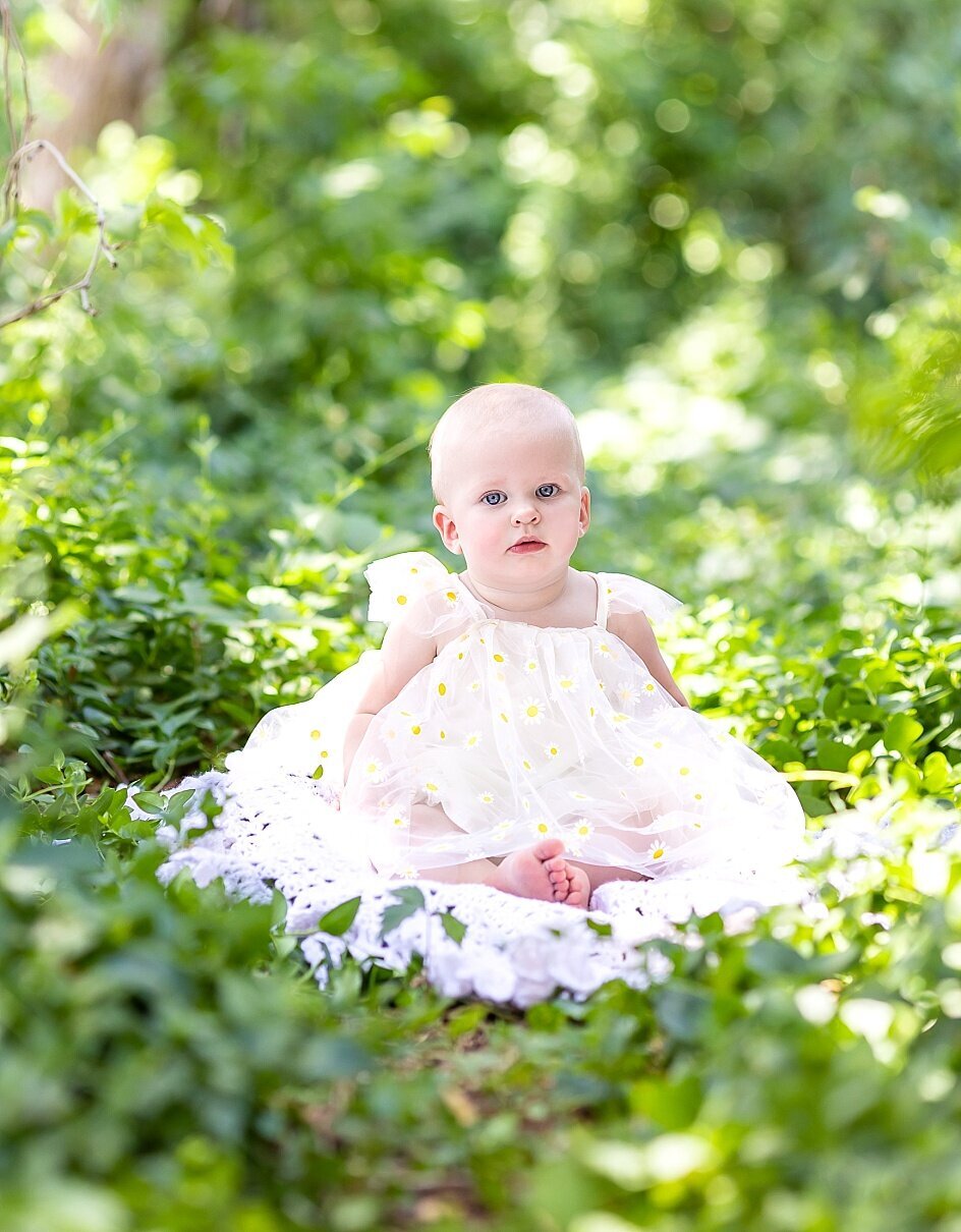 Toddler girl is wearing a white lace dress while sitting amongst plants and looking at the camera