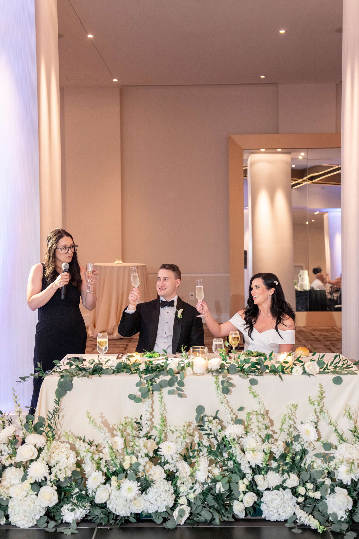 A woman gives a toast with a microphone at a wedding reception. The bride and groom, seated at the floral-decorated table, raise their glasses in response. The room is elegantly lit and decorated with white flowers and greenery.