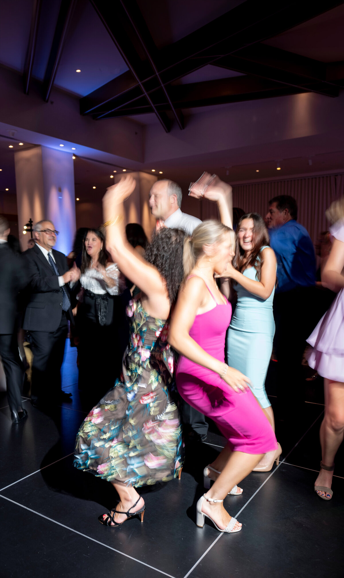 A lively group of people dancing with joy at a party. The scene is filled with motion as they laugh and move energetically on a dimly lit dance floor, surrounded by festive lights and other guests in the background.
