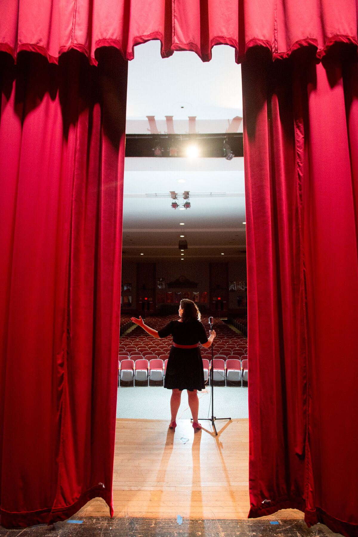 young girl in black dress is seen from the through red stage curtains lit by a spotlight theater kid senior portrait photographer Tappan Zee High School Milestone Images