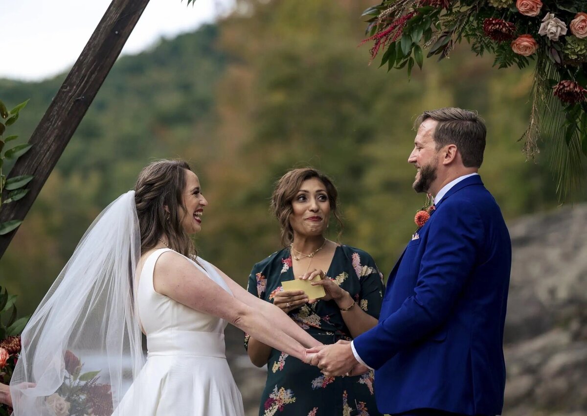 A bride and groom hold hands during their outdoor ceremony, with an officiant between them, against a backdrop of lush greenery and floral arrangements.