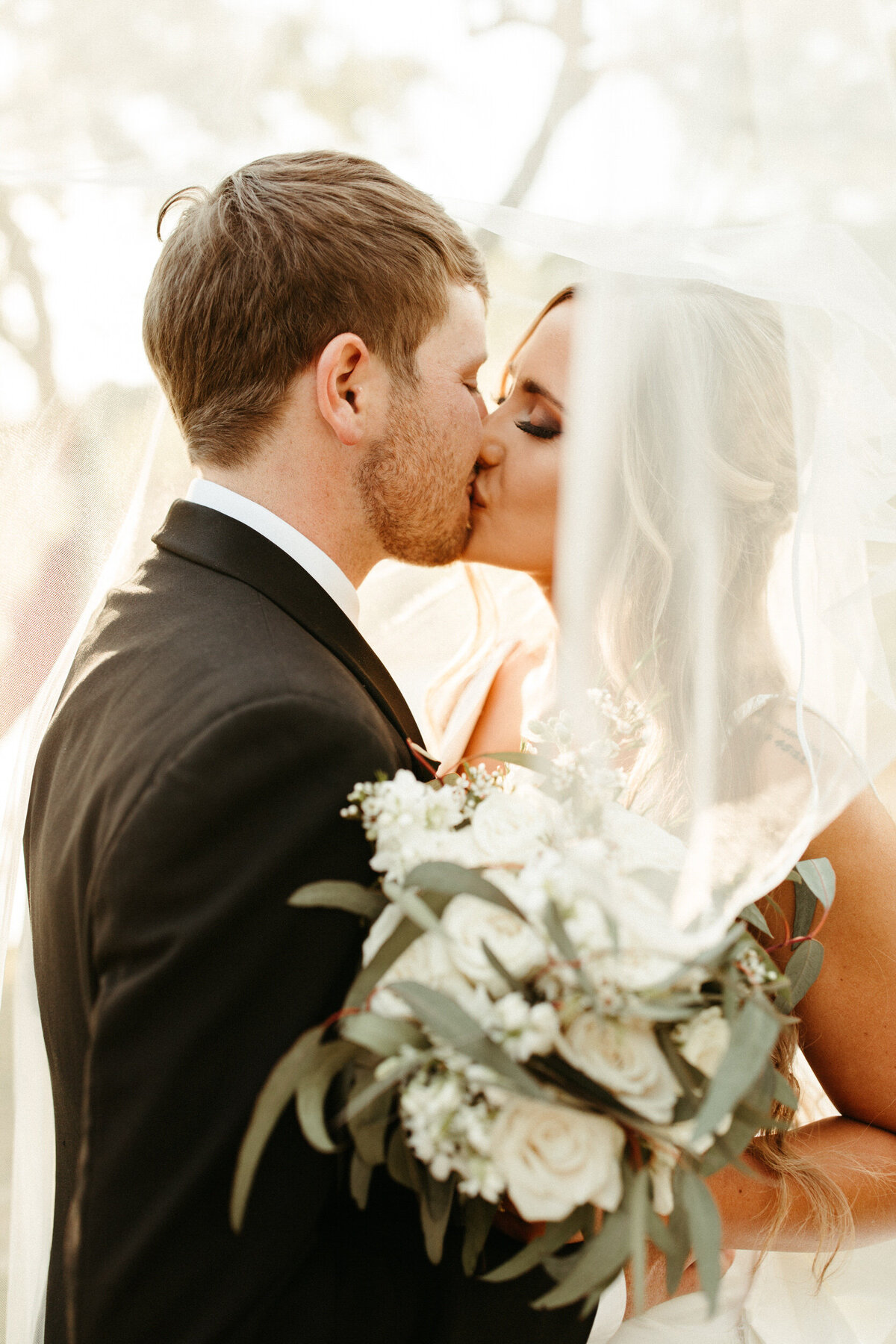 Classic bride and groom kissing underneath her veil as she holds her neutral white bouquet with greenery