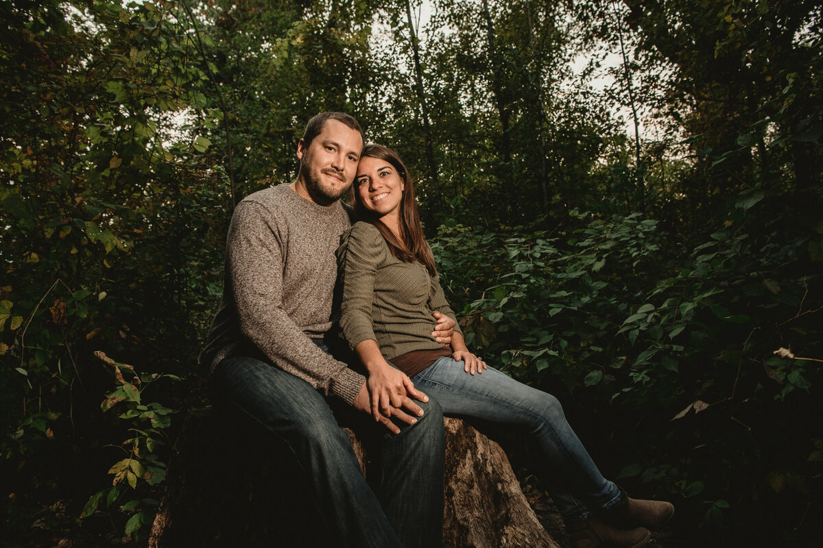 Man and woman in jeans smile on a rock in the woods.