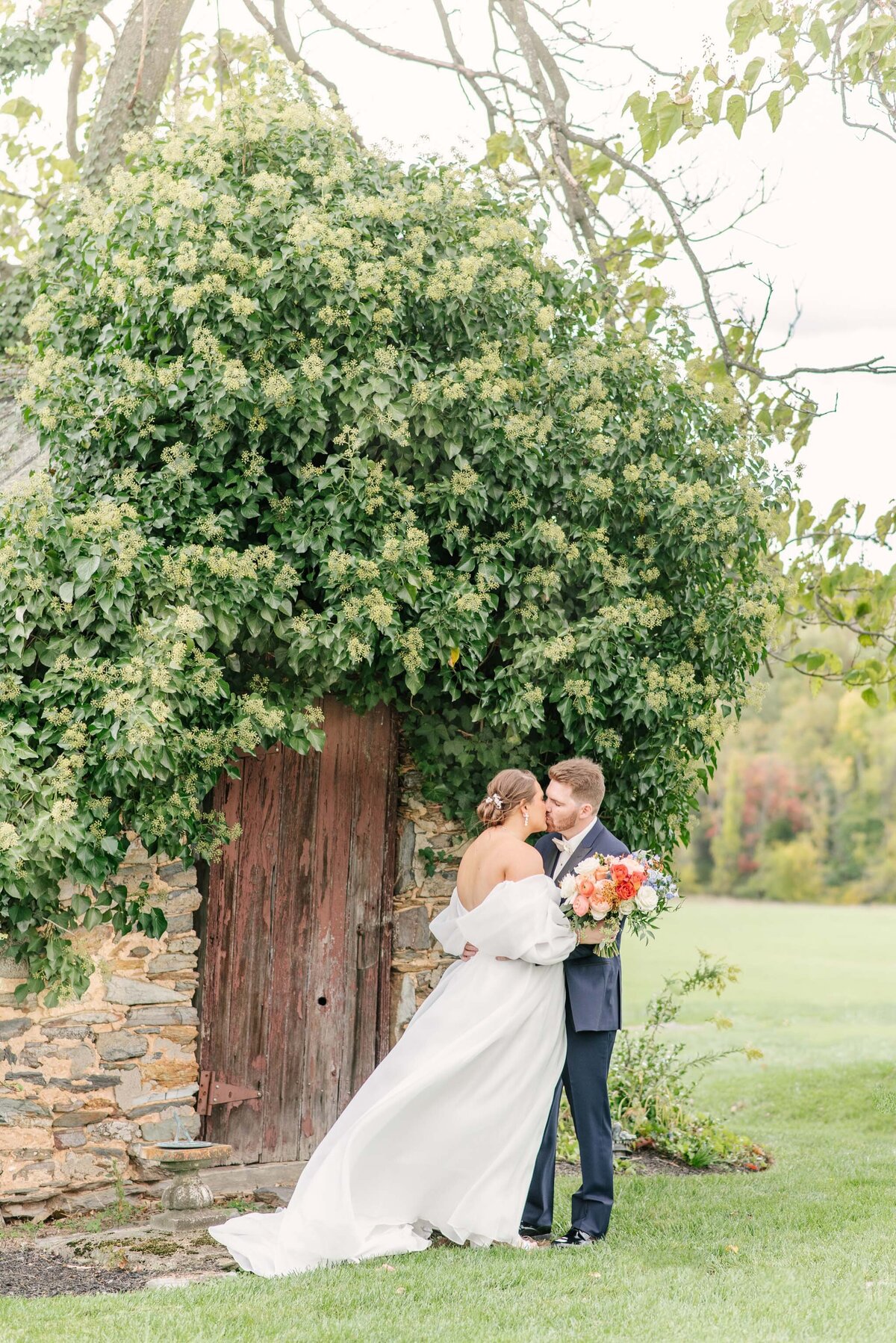couple romantically kissing under greenry and stone wall with orange bouquet and blue suit