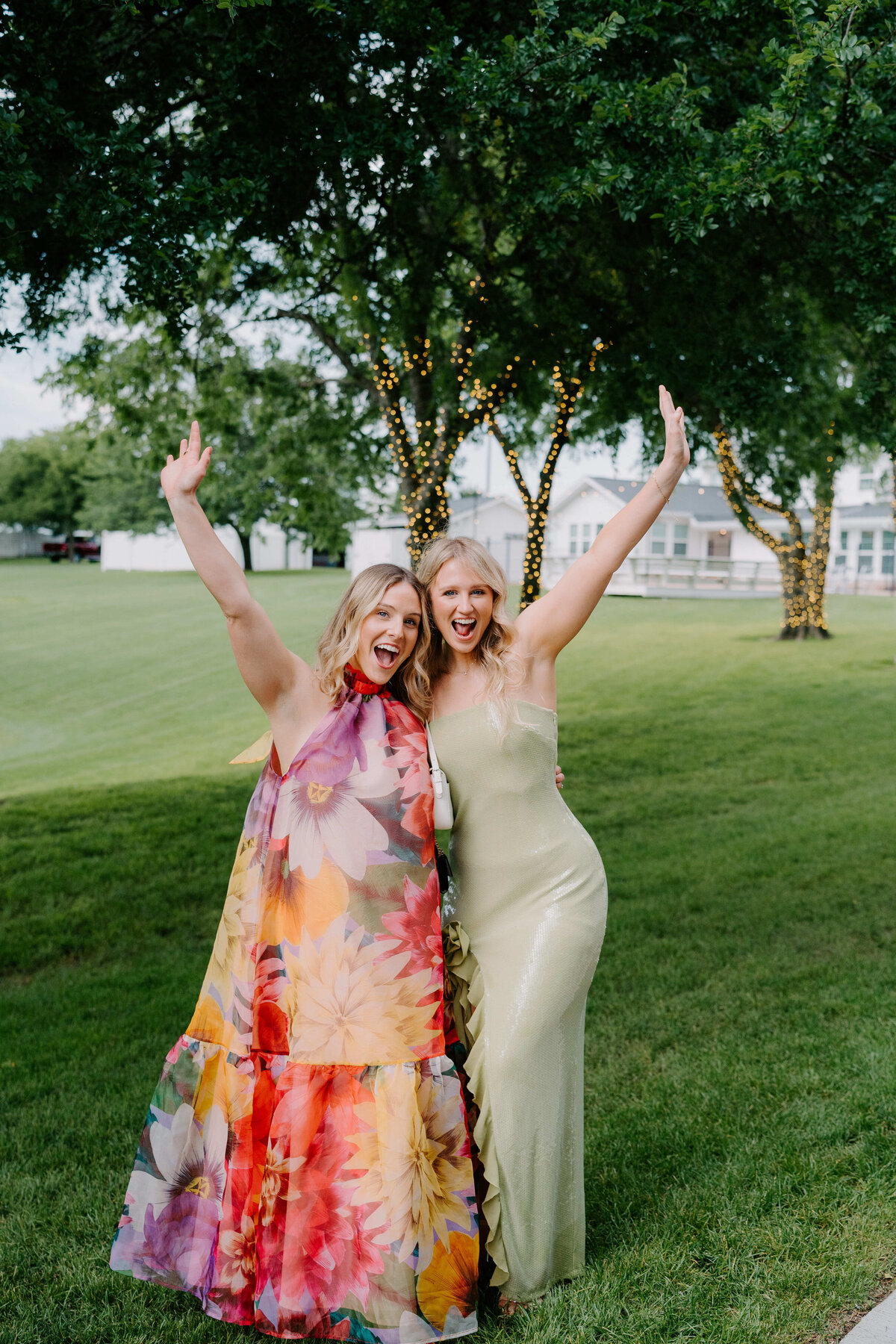 Two women joyfully pose on a grassy lawn. One wears a vibrant floral dress, the other a light green gown. String lights adorn trees in the background.