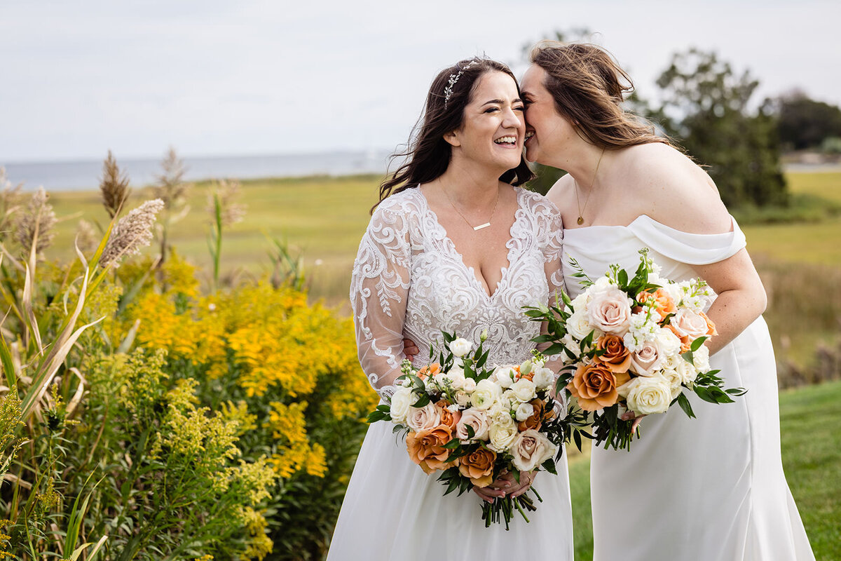 Two brides standing close in a grassy field, one kissing the other on the cheek, both holding bouquets with orange and white flowers, with tall grass and a blue sky in the background.