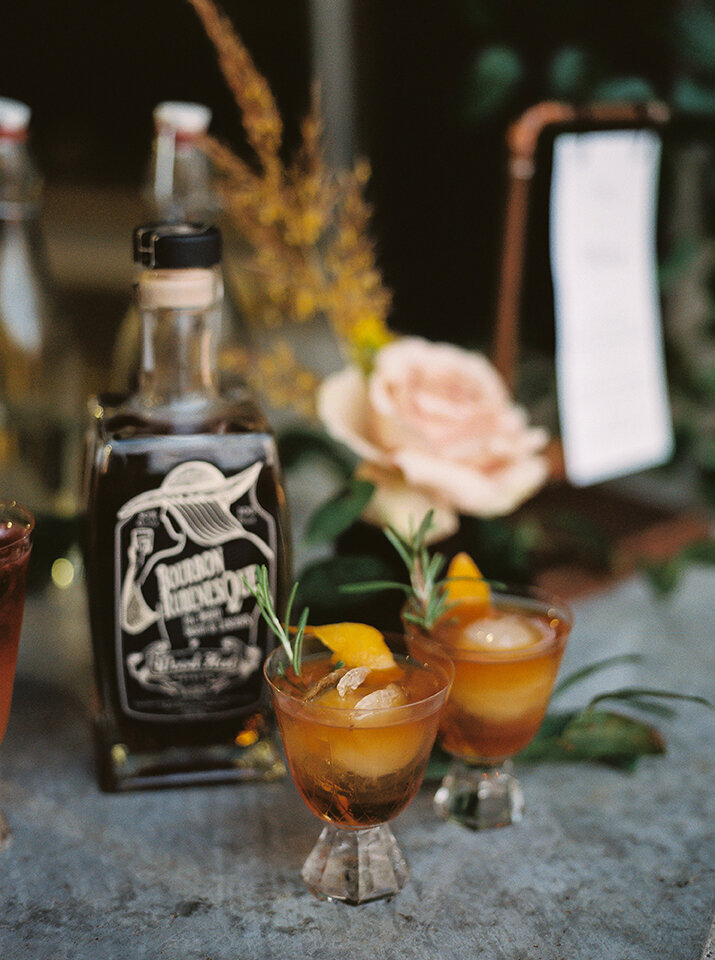 A bottle of bourbon and two cocktails atop a table with blush flowers and a place card.