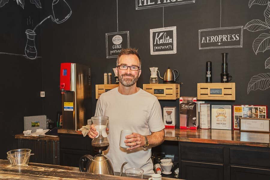A smiling man with glasses stands behind a coffee bar filled with various brewing equipment and coffee bean packages.