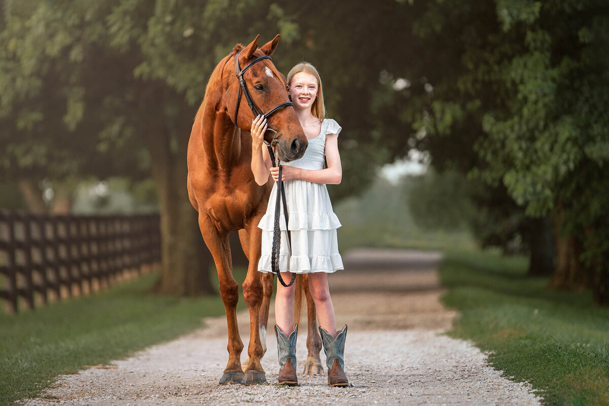 Young blonde equestrian wearing a white dress and cowboy boots is standing next to her chestnut Thoroughbred gelding. they are standing on a gravel path lined with green grass and full green trees behind them.