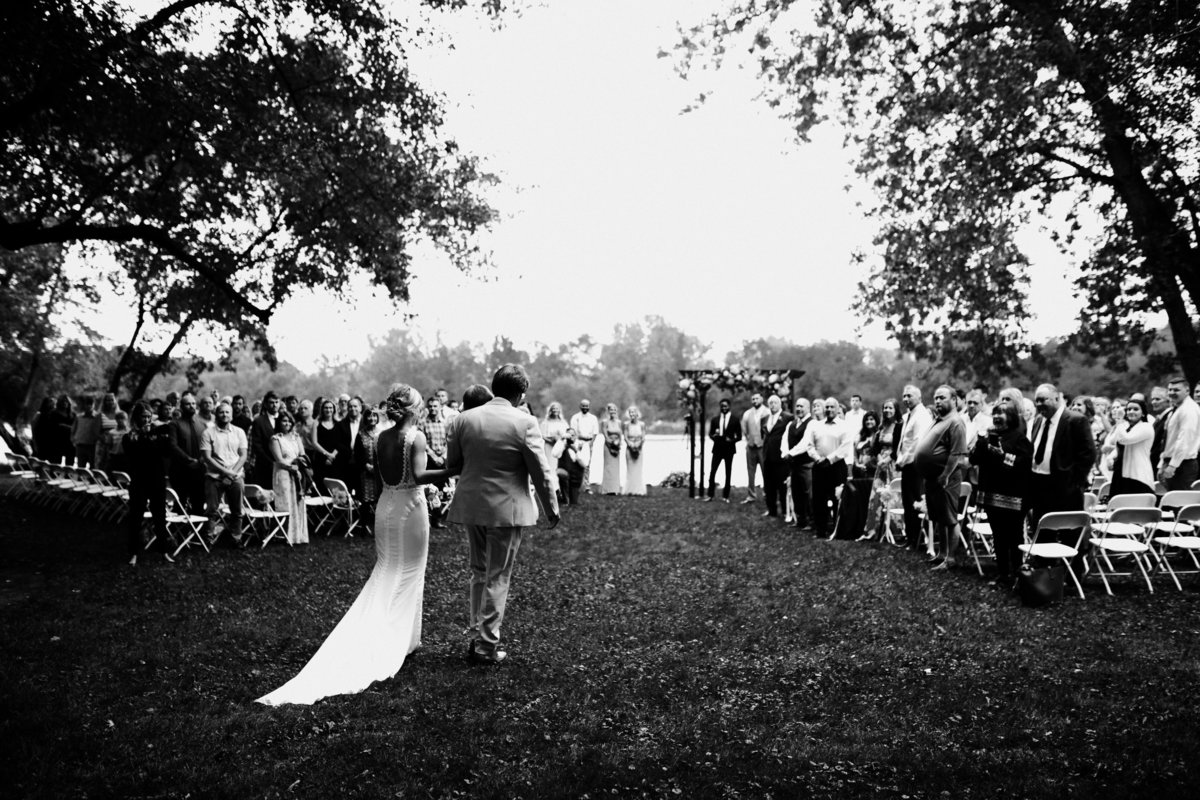One of the top wedding photos of 2020. Taken by Adore Wedding Photography- Toledo, Ohio Wedding Photographers. This photo is of a bride and her father at a wedding ceremony on the bank of a river as guest watch the groom tear up.