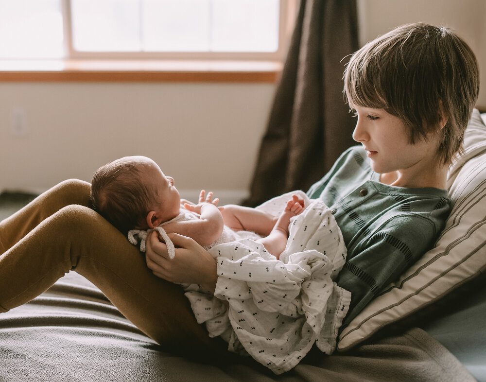Older sibling gently holding newborn on their lap in natural window light, capturing a quiet, connected moment during a baby-led newborn session in Portland.