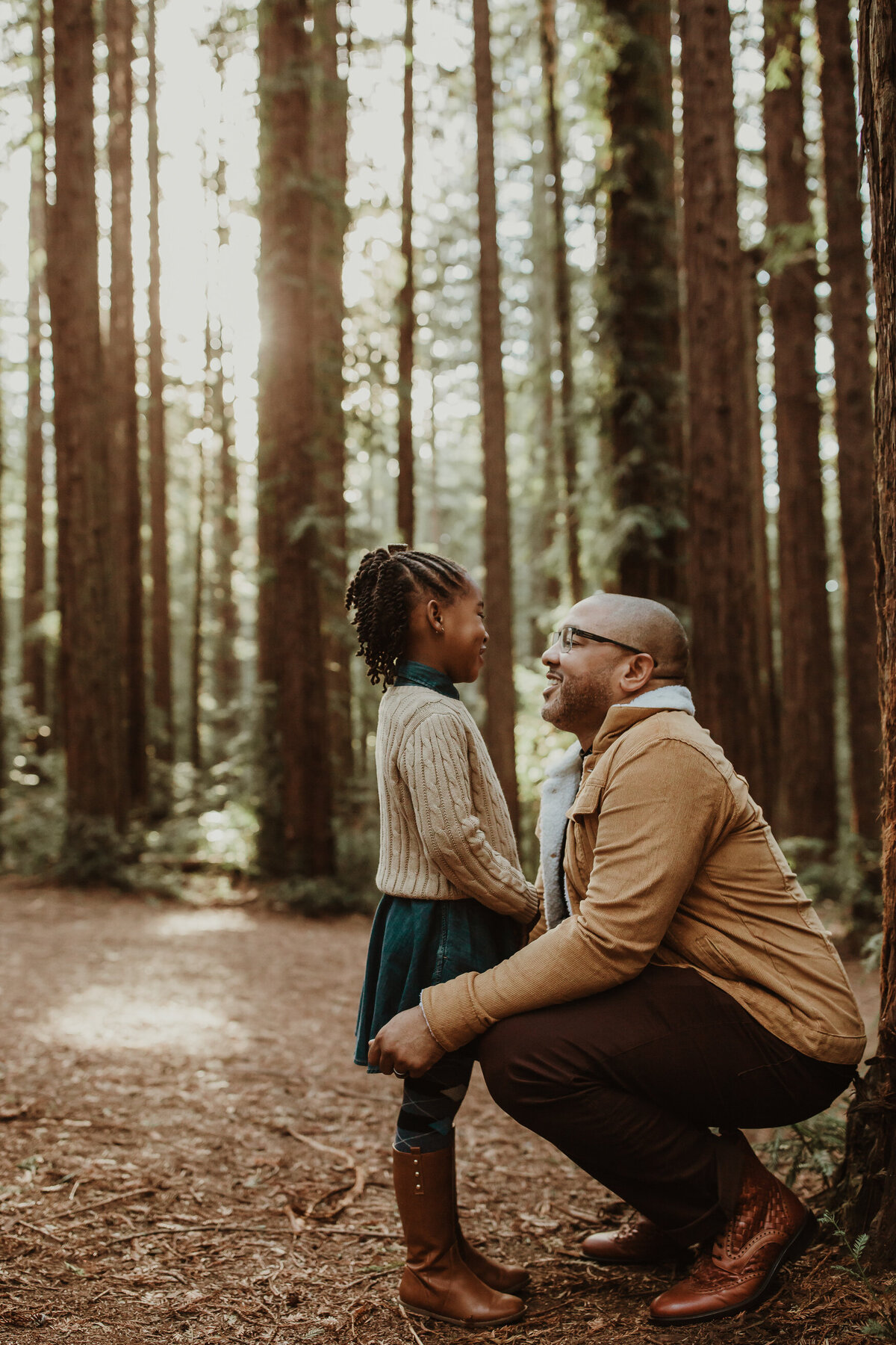 Lifestyle image of father crouched down looking at daughter in Oakland Joaquin Miller Redwood forest.