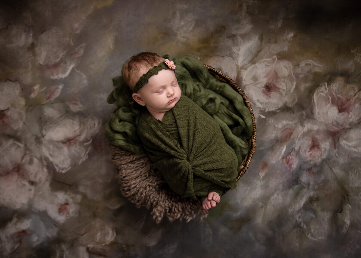 newborn baby asleep in a wooden bowl wrapped in green fabric wearing a floral headband laying on a floral backdrop