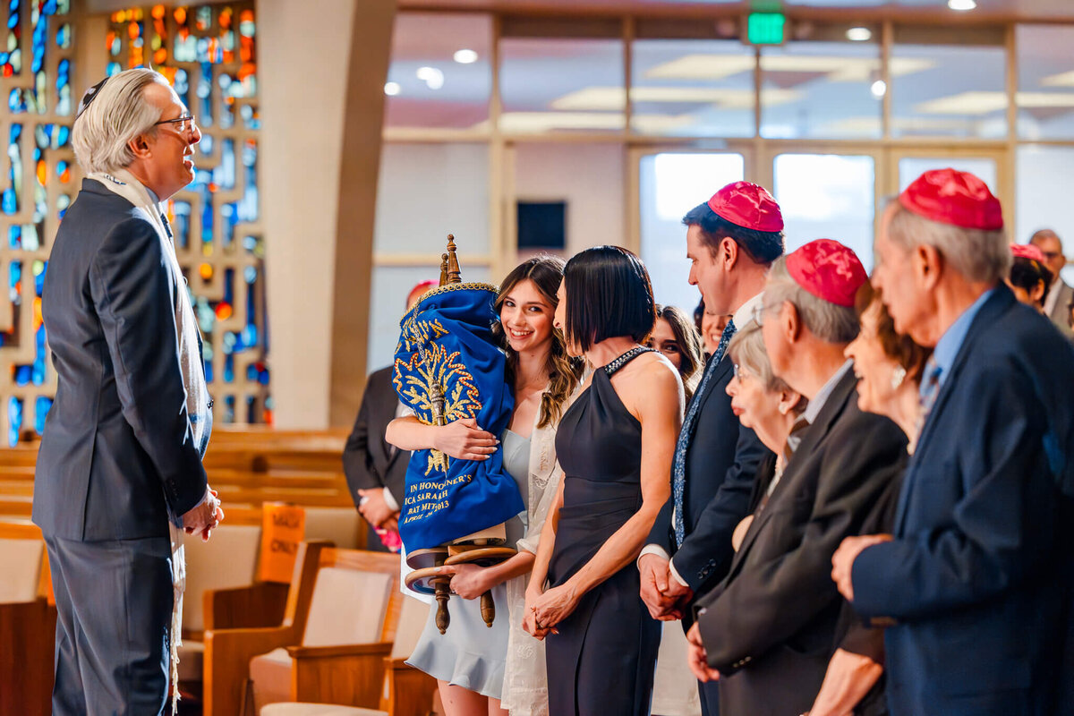 A teenage girl in a blue dress holds the torah smiling with her family and rabbi taken for Bellevue Bar and Bat Mitzvah Photography