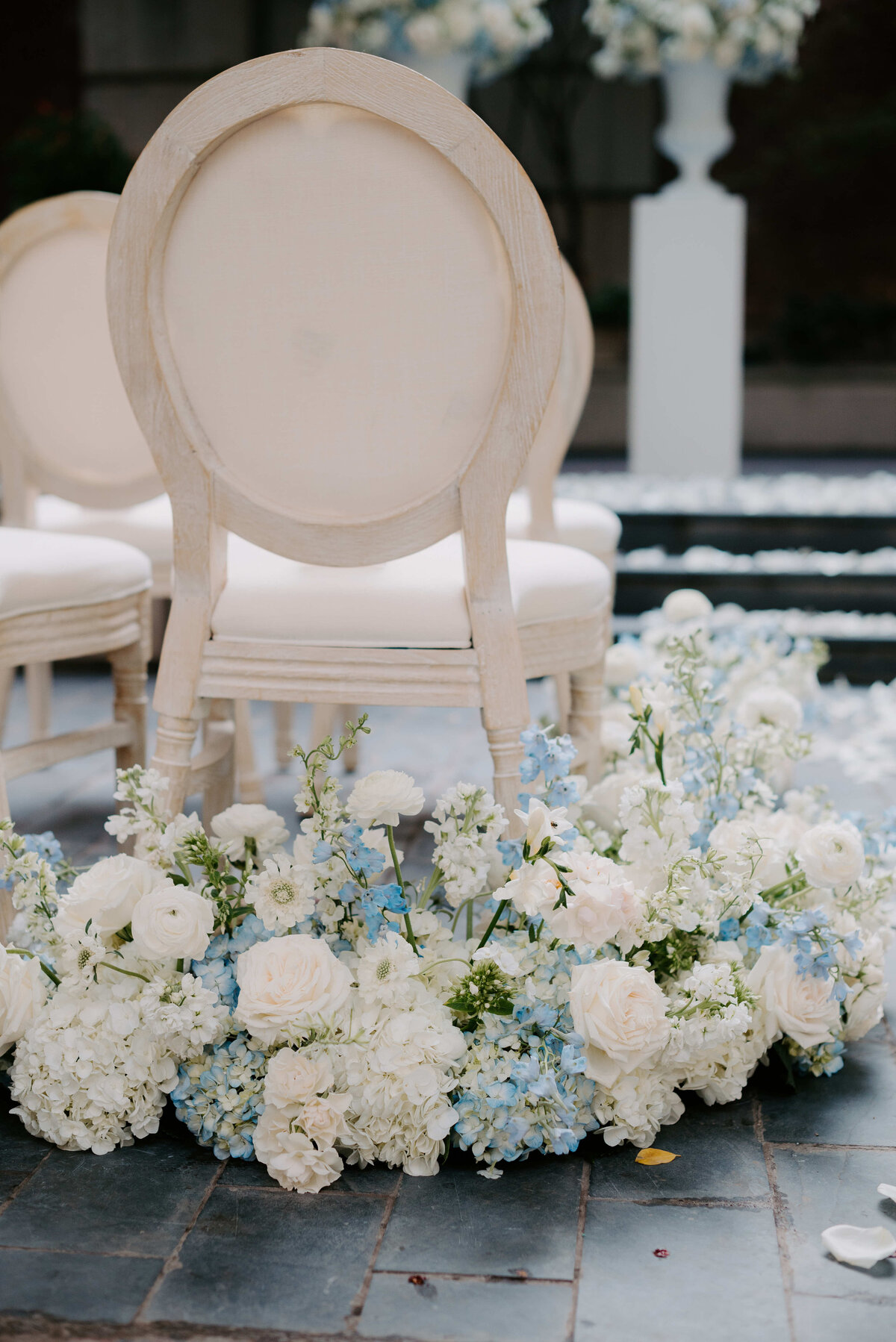 Elegant white chairs are surrounded by lush floral arrangements of white and light blue flowers on a paved path, evoking a serene and romantic atmosphere.