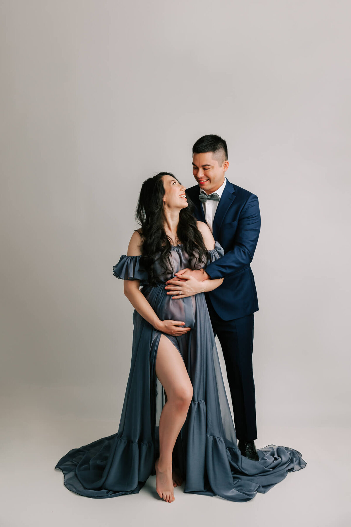 maternity portrait of a couple wearing blue suit and blue dress. She is smiling at him.