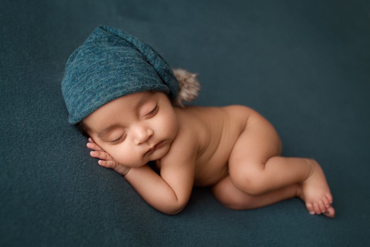 baby boy sleeping on a teal blanket wearing a hat with a pom pom