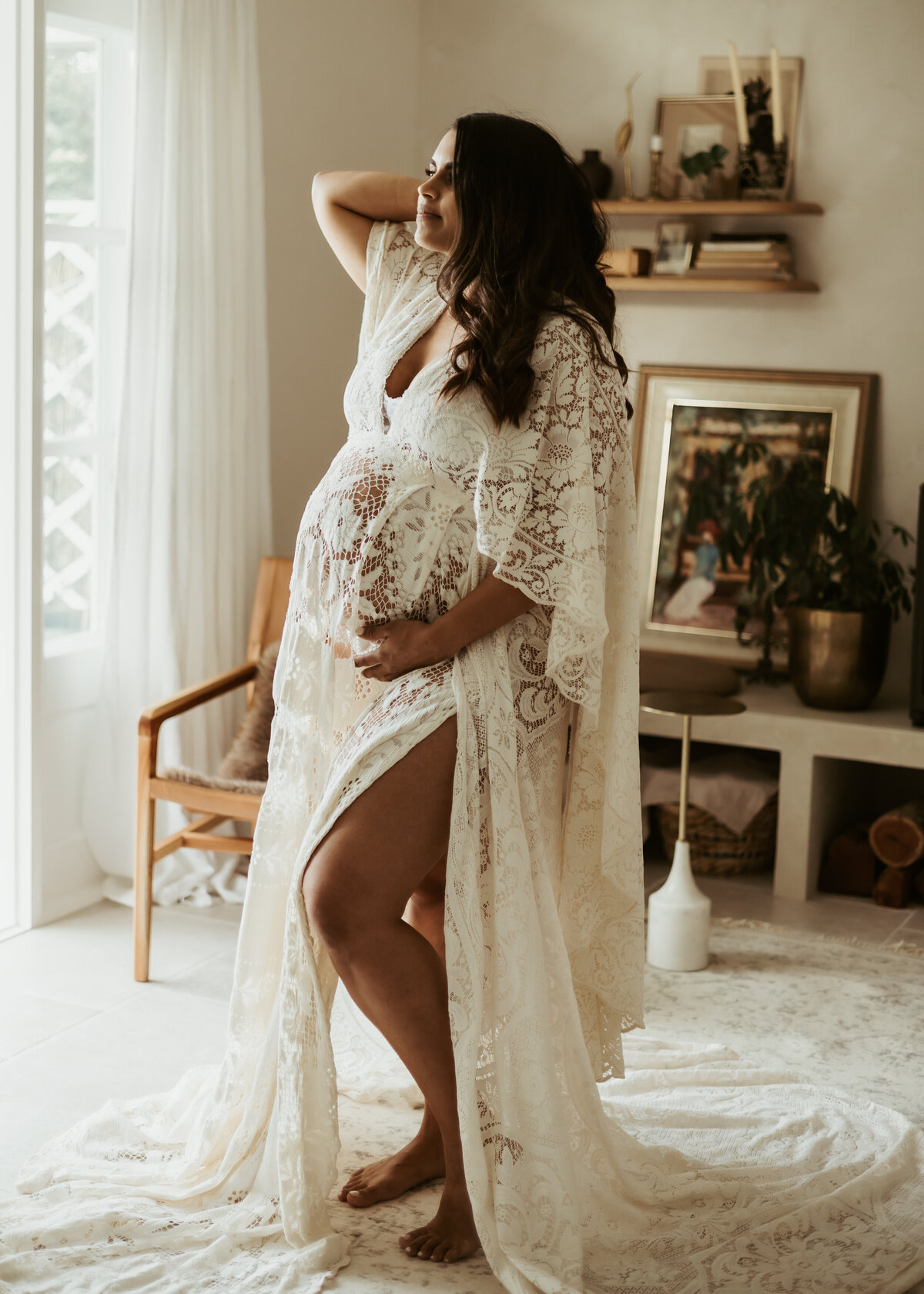 Pregnant mother to be posing for her in-home maternity session in a long white lace dress with slit up the side and holding pregnant belly