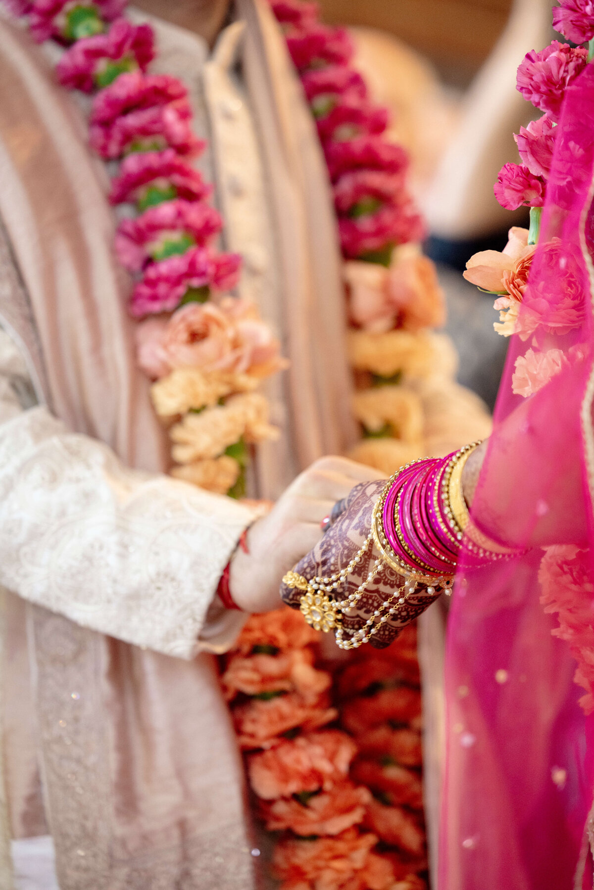 A couple in traditional attire holds hands during a wedding ceremony. They are adorned with layered flower garlands and intricately embroidered clothing. The bride wears vibrant pink bangles and a golden bracelet.