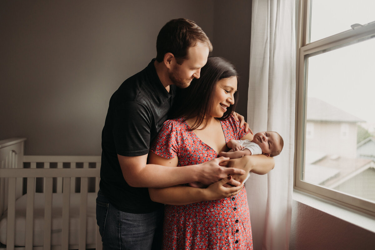 dad wraps arms around mom while holding new baby  infront of  a bright window