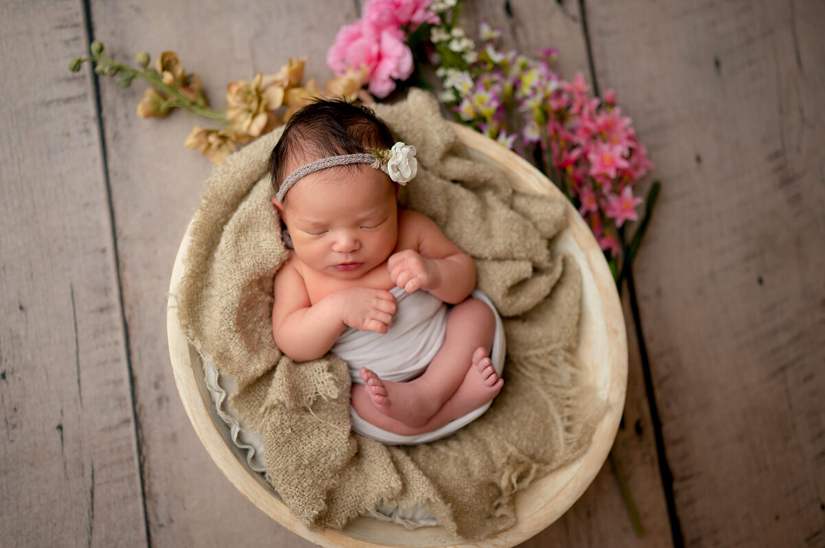 Portrait of a baby girl asleep in a wooden bowl wrapped in cloth. She is flanked by flowers and wearing a floral headband.
