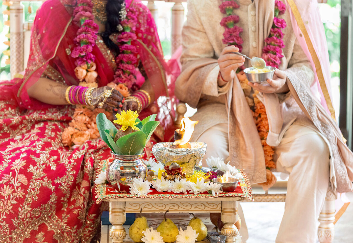 A couple in traditional Indian attire sits at a wedding ceremony. They wear vibrant floral garlands and ornate clothing. An altar with flowers and a small fire is in front of them, signifying a Hindu wedding ritual.