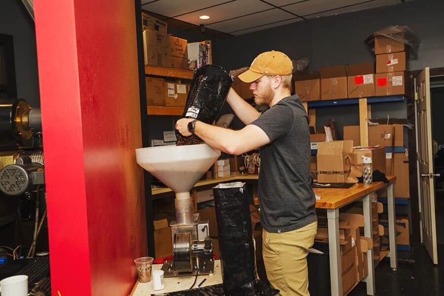 A man in a cap pours coffee beans into a grinder in a storeroom filled with boxes and equipment.