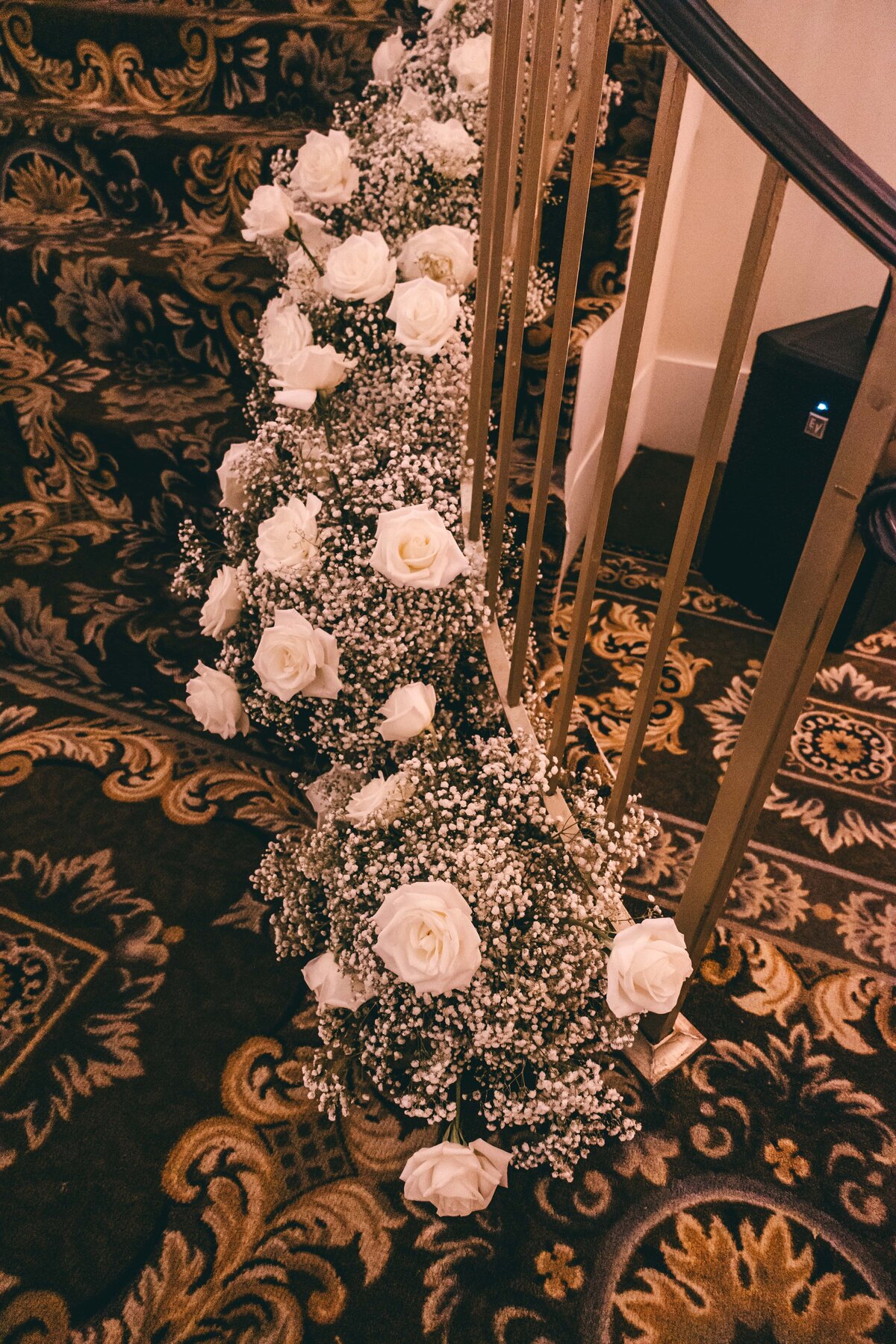 A decorative floral arrangement of white roses and baby's breath along a stair railing, designed by a wedding planner in Des Moines, against a patterned carpet.