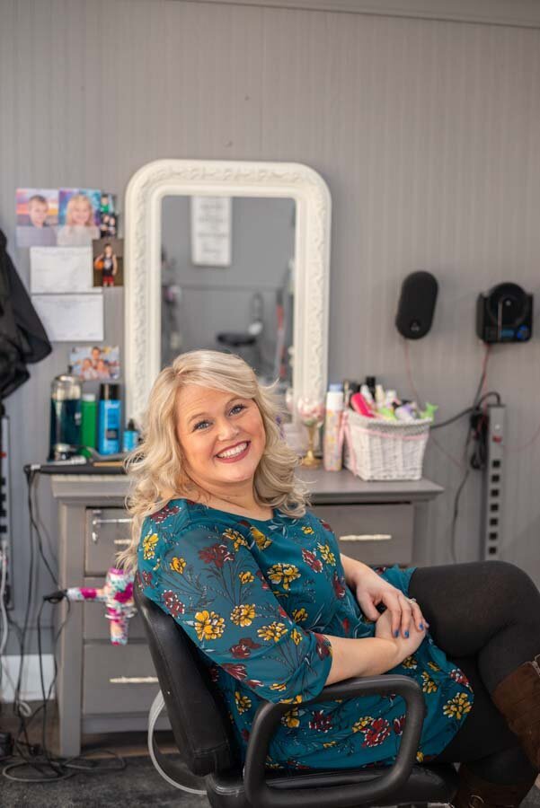 A smiling woman with blonde hair, wearing a floral dress, sitting in a salon chair in front of a mirror and products.
