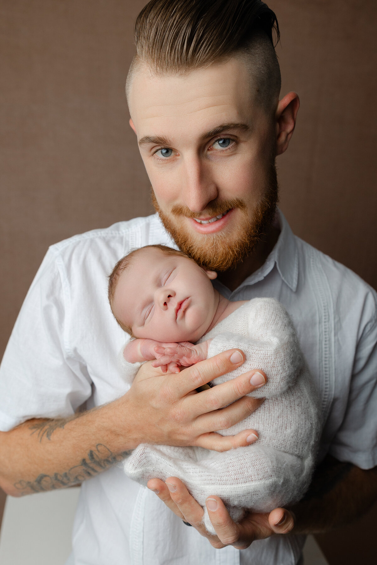 Father smiling while holding his newborn daughter dressed in a white knit outfit during their studio session. Captured by Inland Empire newborn photographer Kaitlyn Dawn Photography.