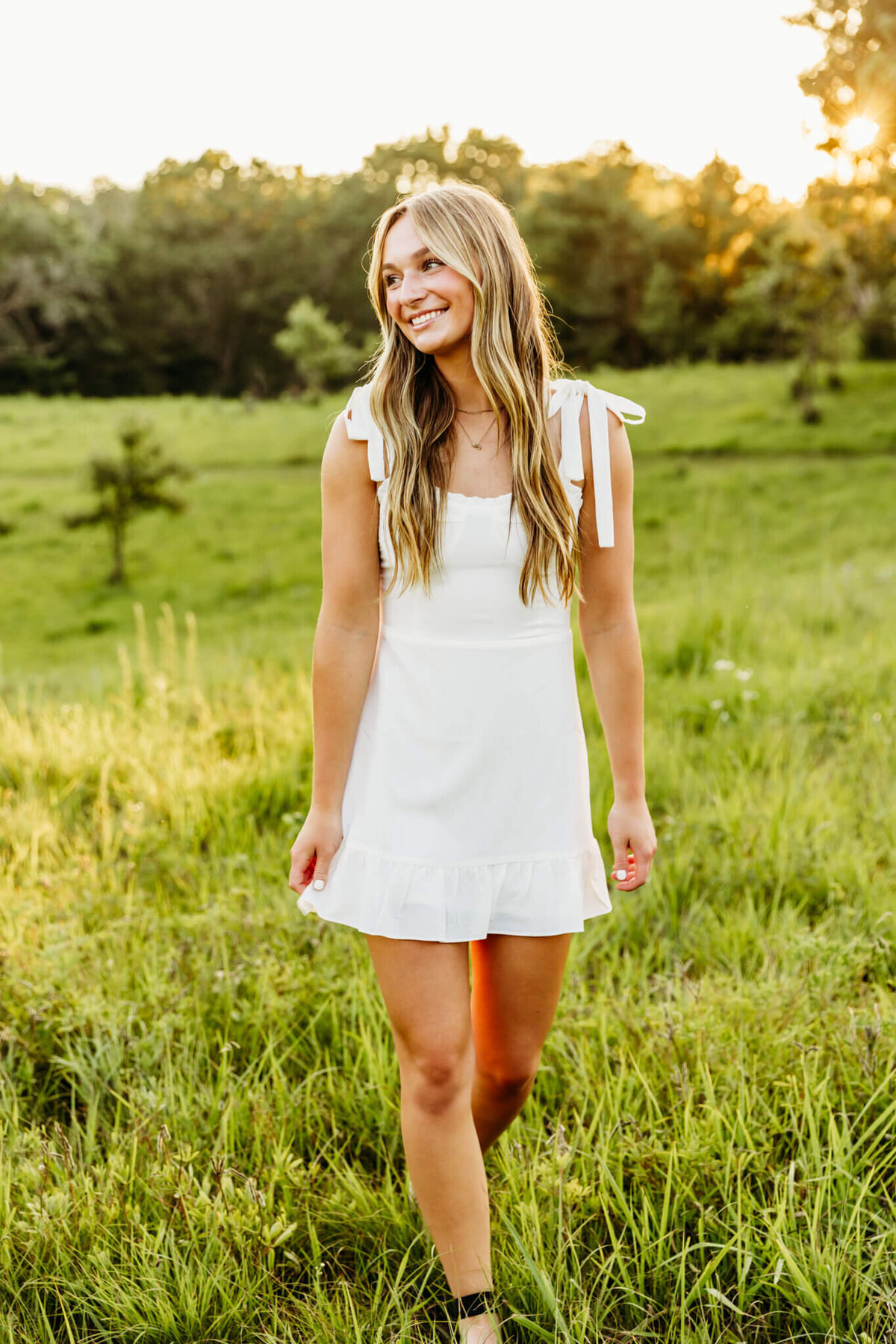 senior girl in an adorable short white dress smiling and looking out in the distance as she walks through a field in Wild Rose