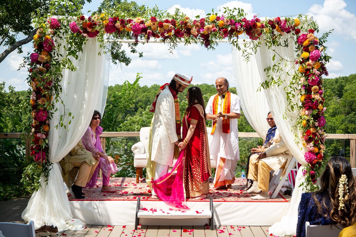 A bride and groom perform a ritual under a floral arch at an outdoor wedding in Iowa, with guests seated around them.