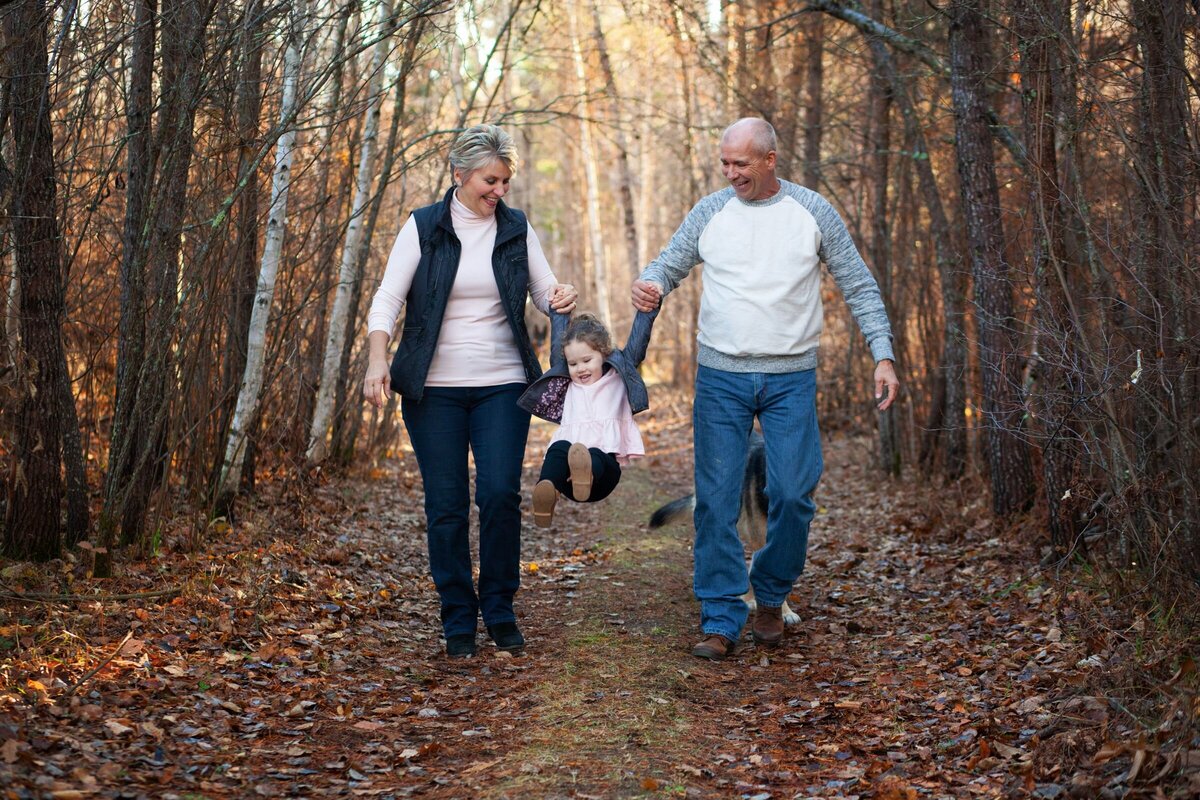 Grandma and Grandpa enjoying a moment with their granddaughter by swinging  her in sync walking down a woodsy path.