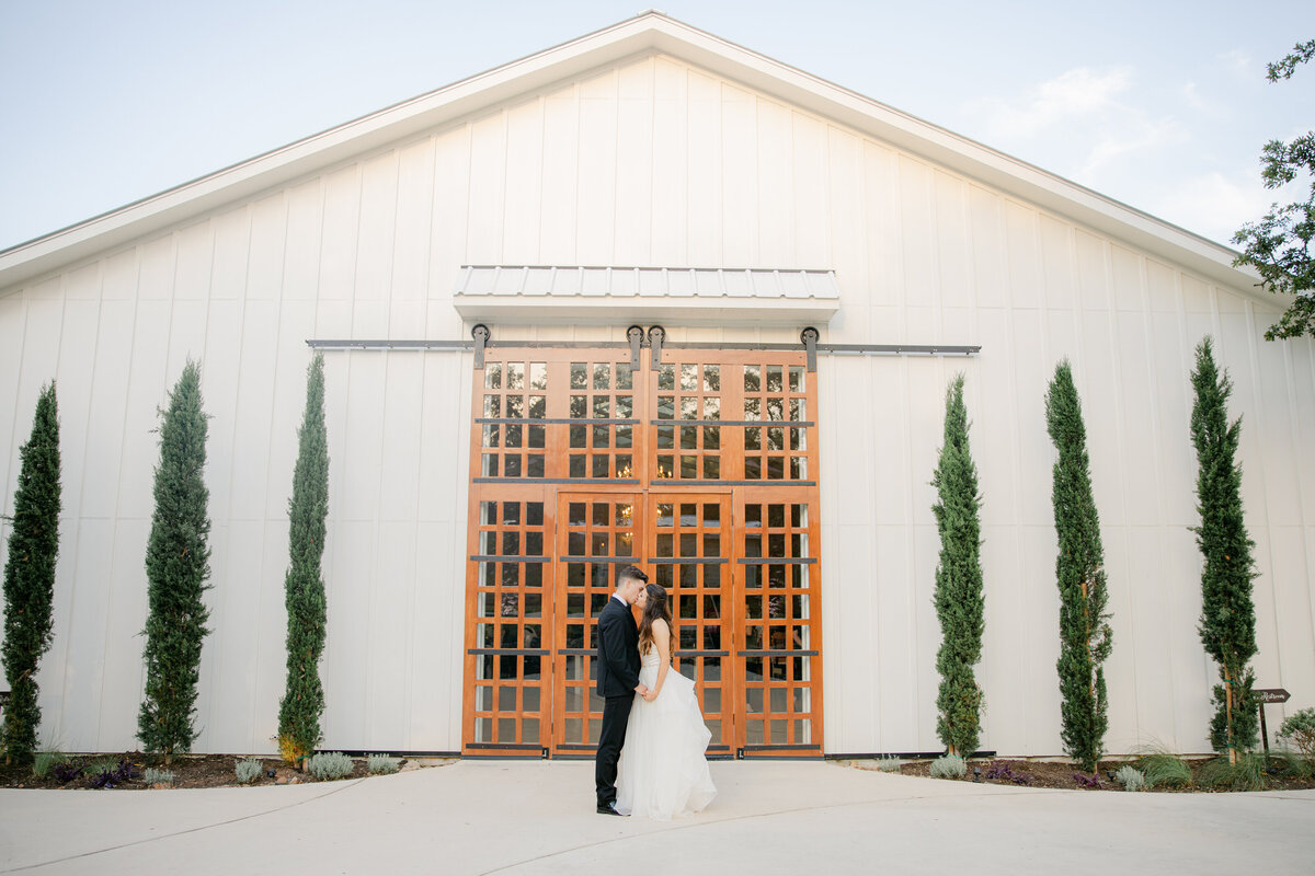 a bride and groom facing each other and standing in front of wooden doors and tall trees in texas