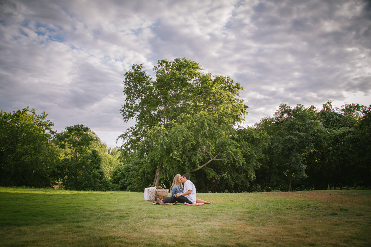 Engaged couple sitting and having a picnic in an open field with a lot of clouds in the sky in Boerne.a