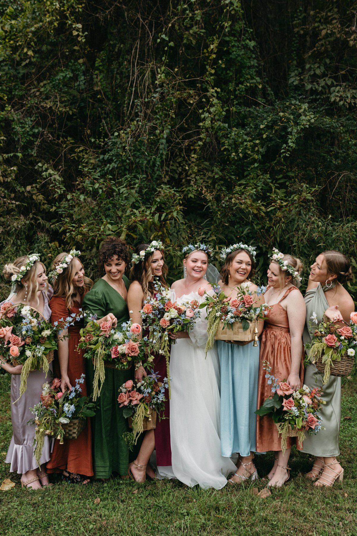 Bridal party grouped together outdoors with lush woodland greenery backdrop, bride in a soft organza gown holding a garden bouquet, surrounded by bridesmaids in mismatched dresses with floral crowns and overflowing basket arrangements.