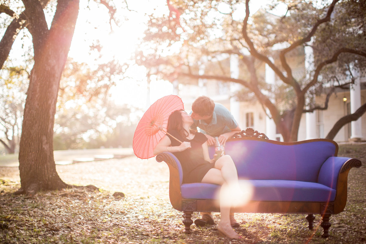 Engagement photography session of couple on a couch kissing with sunlight coming through the trees at Denman Estate Park in San Antonio, Texas.