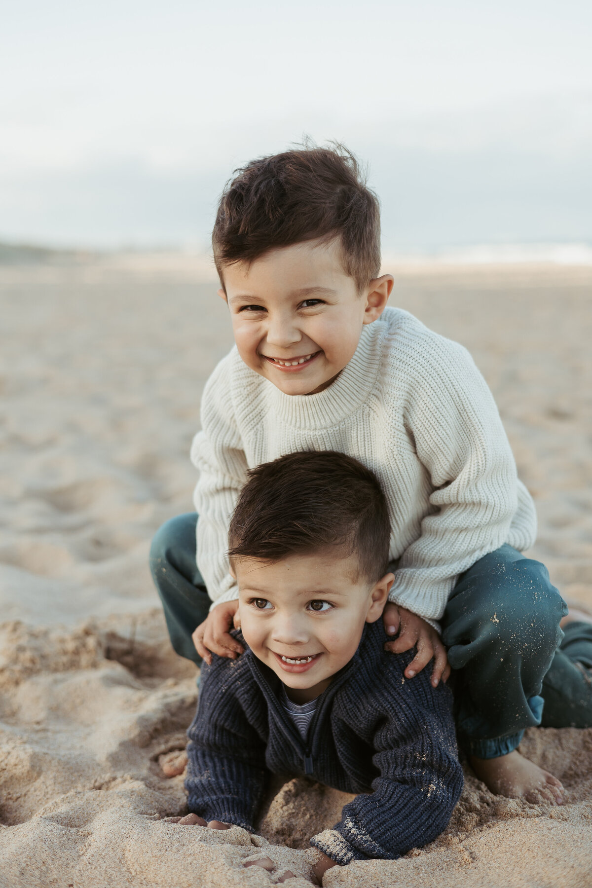 Little brother is laying in the sand while his brother sits on him and smiles for the camera.