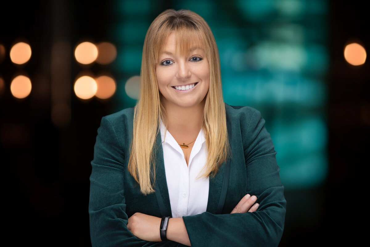 Professional headshot of a smiling Caucasian woman with blonde hair and bangs, wearing a green blazer over a white blouse. Captured in a Cincinnati studio, the background features a soft bokeh effect with warm lighting, highlighting her friendly and approachable demeanor. Perfect for business professionals seeking to update their LinkedIn profile or corporate website. This high-quality image showcases expert posing and professional attire, making it an ideal choice for enhancing personal branding and professional presence online.