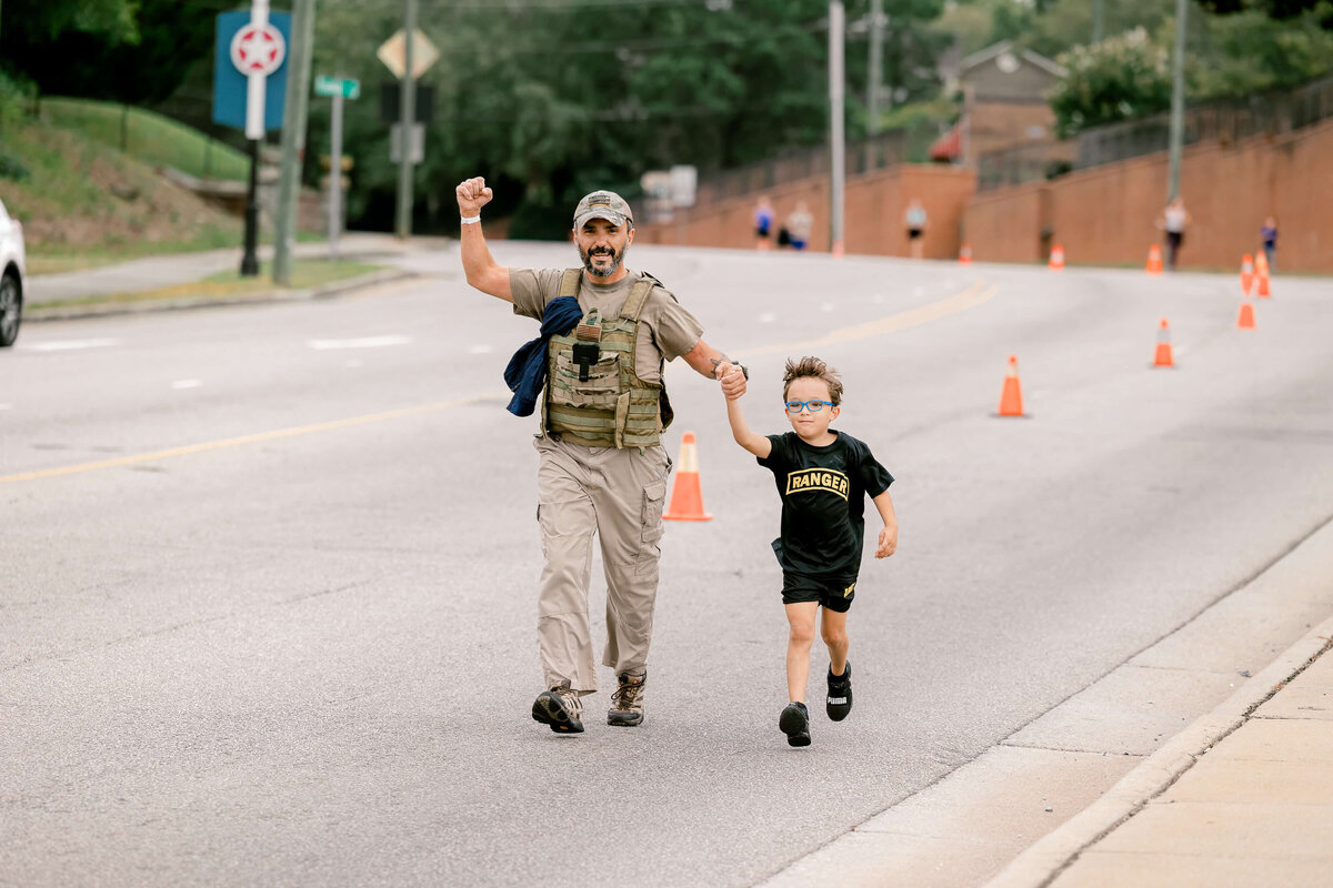 A man and a child dash along a road lined with orange cones. He sports a tactical vest and hat, grasping the child's hand while raising his other arm. The child, in a black t-shirt and blue sunglasses, beams with joy. Captured perfectly by an event photographer from Fayetteville, NC—trees and traffic signs stand behind them.
