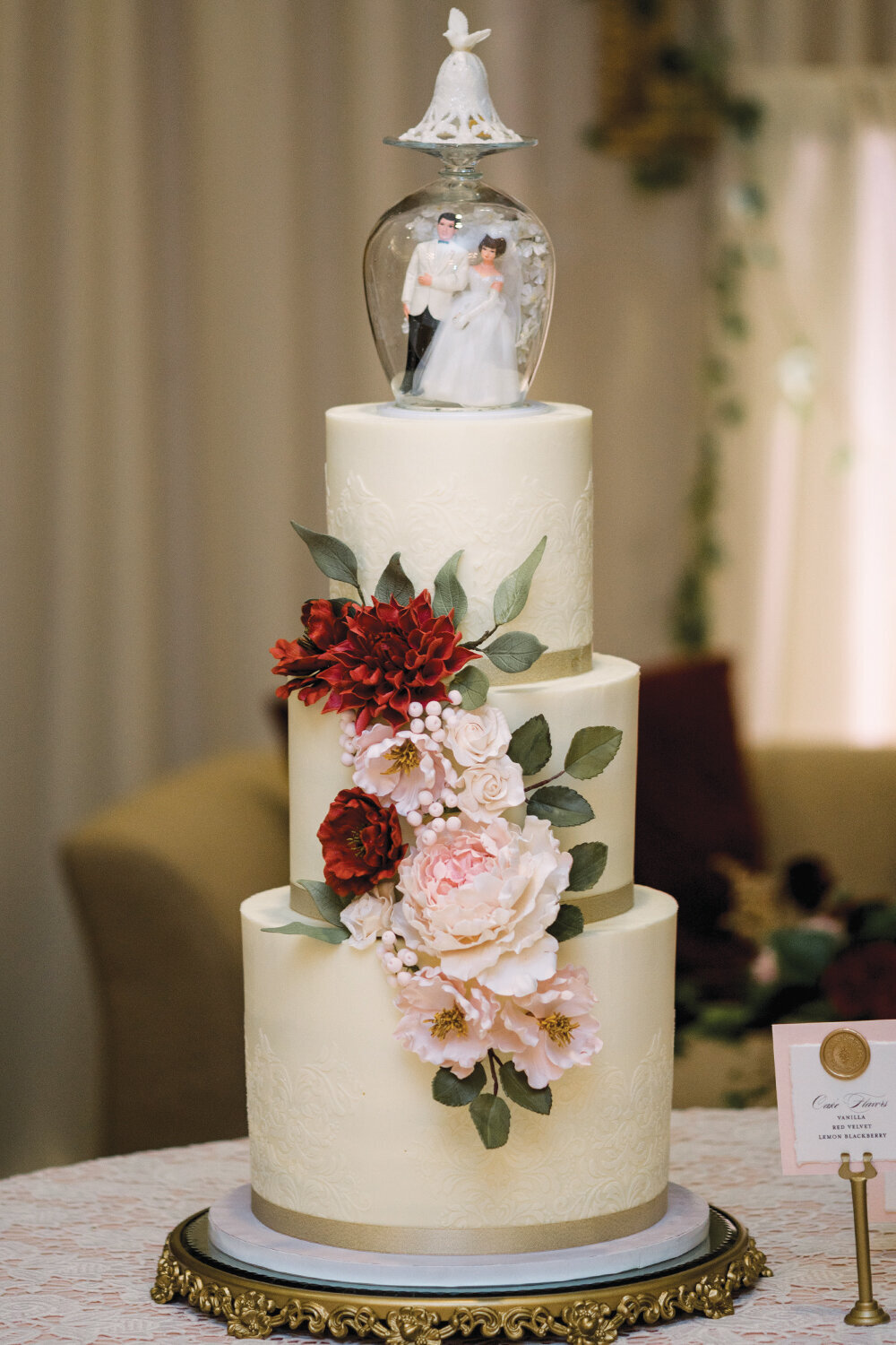 Three-tier wedding cake with intricate lace buttercream detailing, adorned with cascading sugar flowers in burgundy and blush, displayed on an ornate gold cake stand for a 50th anniversary celebration at The Northern Hotel in Billings, MT.