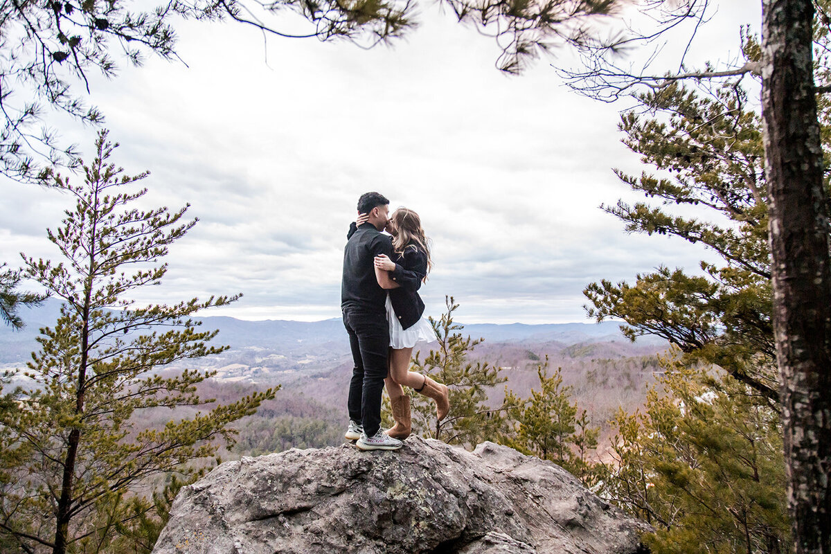 couple kissing on top of a rock with the mountains in the background