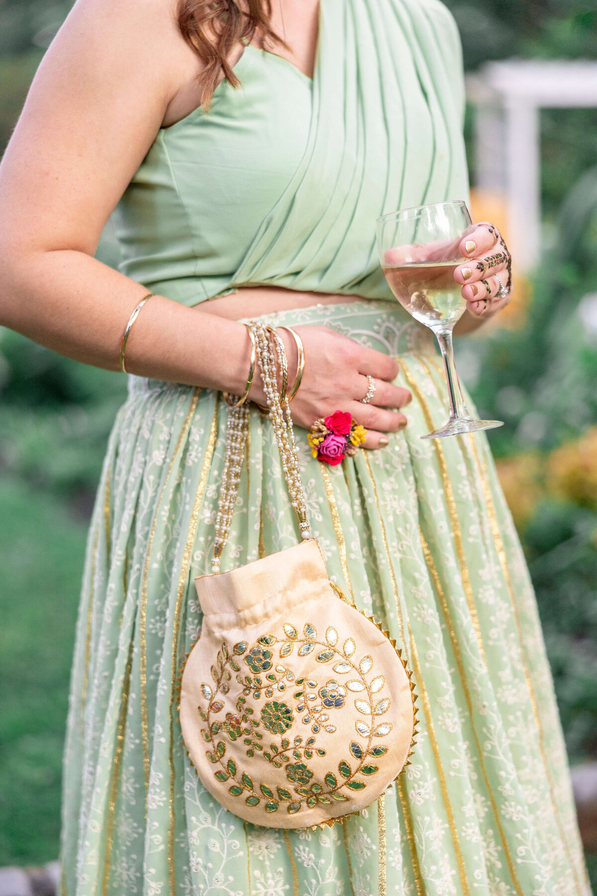 A woman wearing a light green, embroidered two-piece outfit holds a glass of white wine and a beige embroidered purse. She accessorizes with gold bangles and a floral ring. The background is a garden setting.