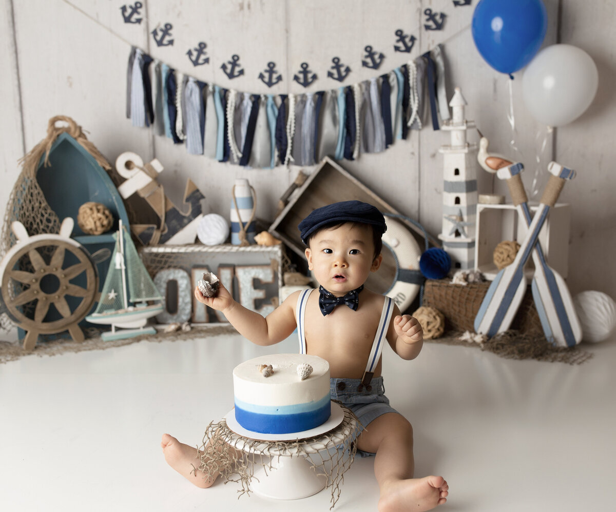 Nautical themed cake smash at West Palm Beach, FL photography studio.  Baby boy is looking at the camera and the ombre blue cake untouched. In the background there are sailboat, blue oars, and lighthouses.