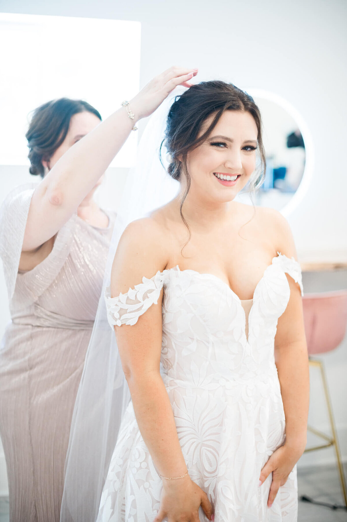 bride shyly smiles at camera while her mother puts in a veil