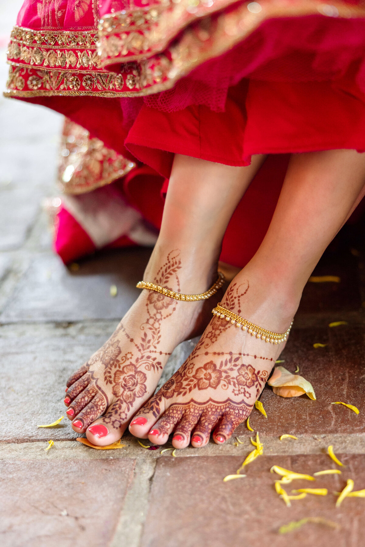 Close-up of a brides feet adorned with intricate henna designs and gold anklets. She wears a traditional red bridal outfit, with yellow petals scattered on the ground beneath her.