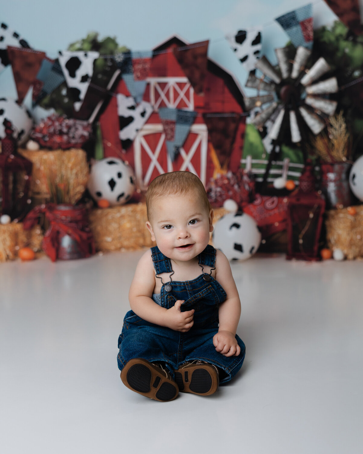 Smiling baby boy sitting on the studio floor in front of a barnyard-themed cake smash backdrop with hay bales and cow print balloons.
Photograph by Yucaipa cake smash photographer Kaitlyn Dawn Photography.