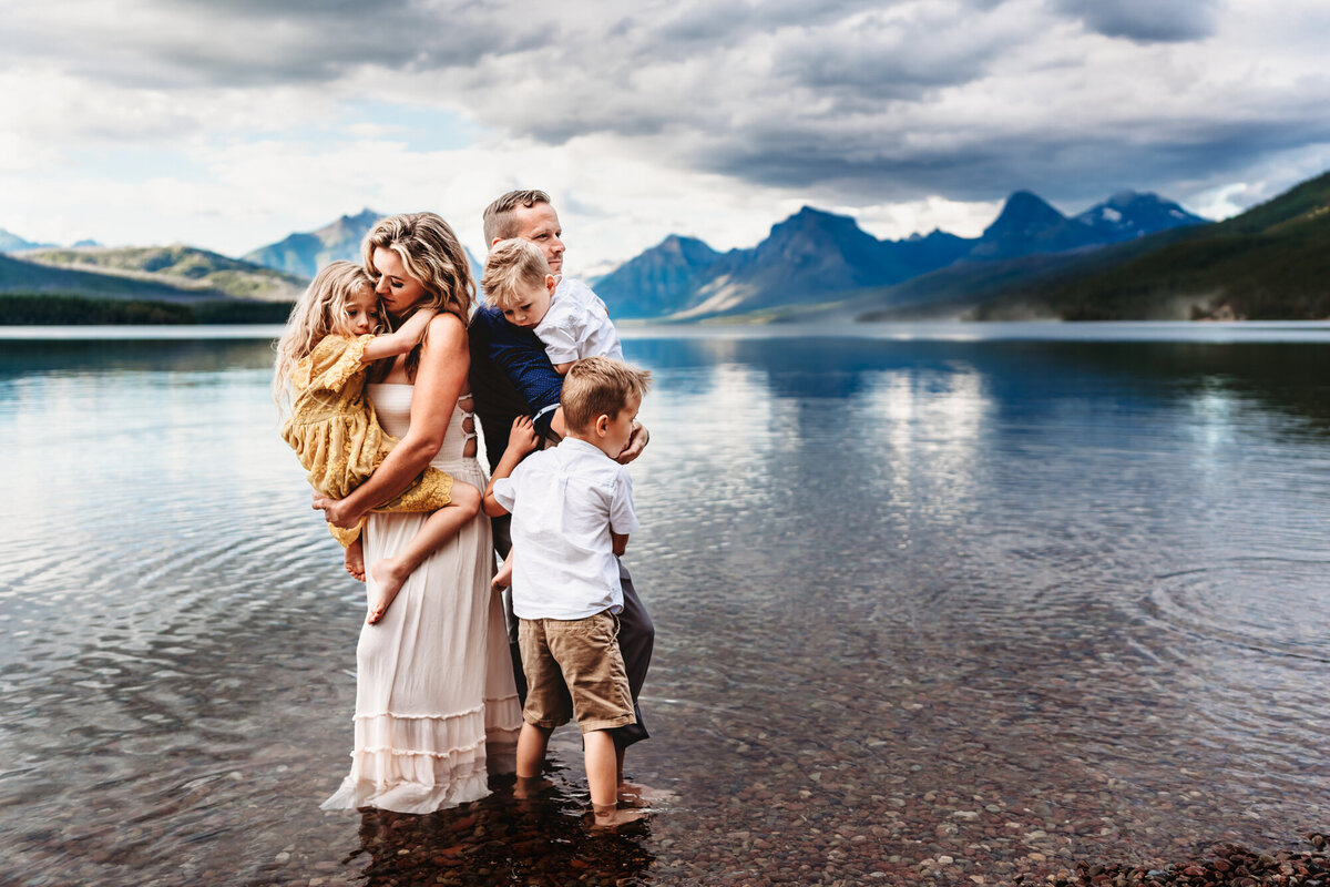 A family standing together on the shores of Lake McDonald in Montana's Glacier National Park,  embracing one another. Taken during a lifestyle vacation family photo session by Love Michelle Photography